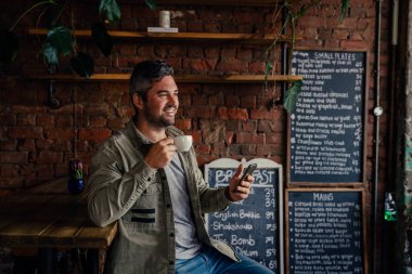 Gorgeous young male grinning while sipping warm coffee at a trendy coffee shop with smartphone in hand 