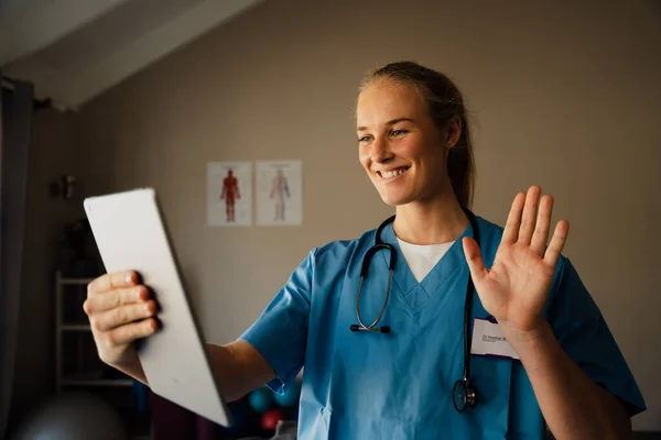 Όμορφη νεαρή καυκάσια γυναίκα γιατρός σε βιντεοκλήση από την πρακτική στο σπίτι, χαμογελώντας και χαιρετώντας τον ασθενή — Φωτογραφία Αρχείου