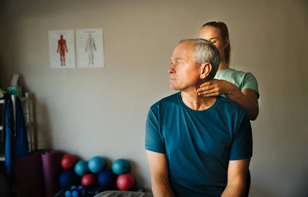 Male patient twisting his neck while female physiotherapist massages pinched nerves of neck standing in rehab room.