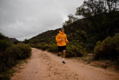 Strong young male athlete completing steep long distance run on gravel mountain path with bushes and trees in cloudy weather