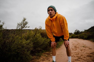 Close up tired young male athlete resting to catch breath after downhill sprinting on gravel trail in cloudy weather with foliage