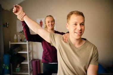 Young female caucasian physiotherapist stretching shoulder of handsome male patient in studio.