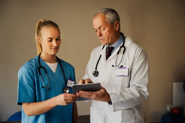 Male doctor teaching female intern in scrubs, while showing instructions on digital tablet in organised doctors room — Stockfoto