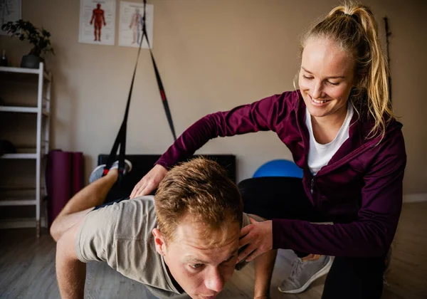 Caucasian female physiotherapist guides male patient doing pushups with black rubber band in exercise studio. — Stockfoto
