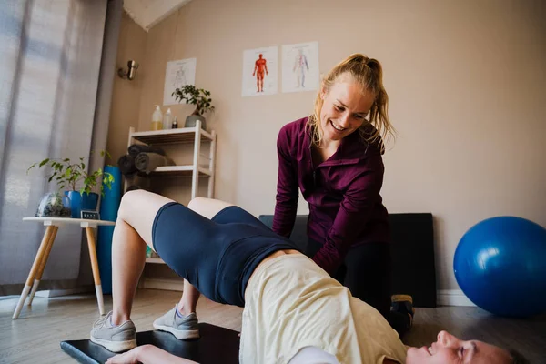 young female patient holding bridge position as beautiful female physiotherapist corrects alignment in exercise studio.