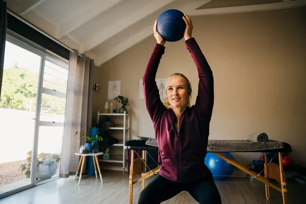 Strong caucasian female squatting holding blue ball in light studio. — Stockfoto