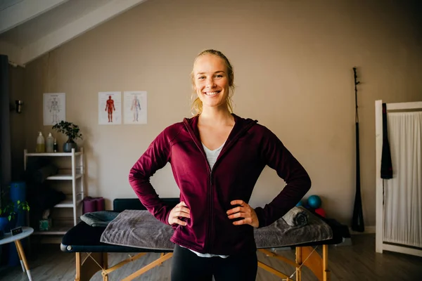 Blonde caucasian female physio standing with hands on hips in front of bed in organised studio. — Stockfoto