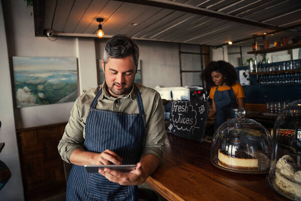 Male waiter scrolling on digital tablet searching for menu while waitress makes coffee in coffee restaurant