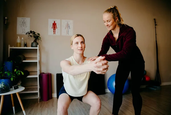 Young female patient practising squat position while trained physiotherapist corrects posture in trendy exercise studio. — Stockfoto