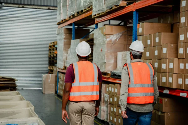 Two mixed race working together walking through isles of packaged parcels in factory — Stockfoto