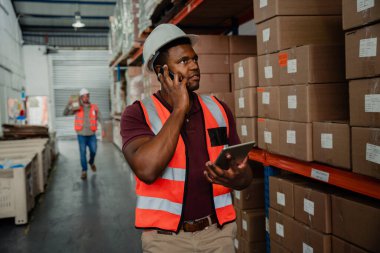 Mixed race male worker chatting to boss on cellular device while holding digital tablet standing in factory 