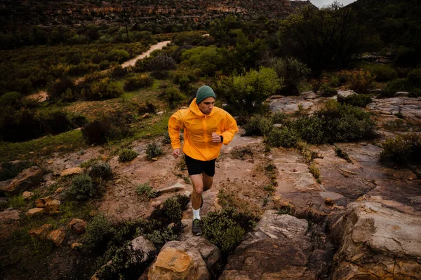 Wide angle fit young male trail runner sprinting up wet rocks on side of mountain with green bushes and hills