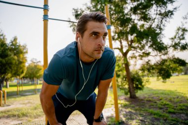 Odaklanmış genç erkek sporcu uzanıp kulaklıklarla müzik dinliyor. Açık hava parkında spor yapıyor.