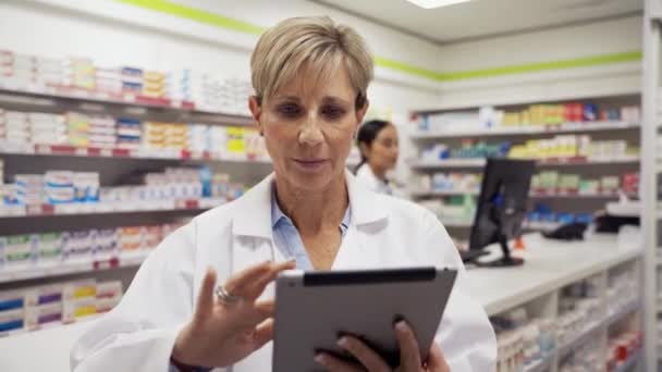 Female pharmacist scrolling through emails on digital tablet while assistant collects medicine behind counter in pharmacy — Stock Video