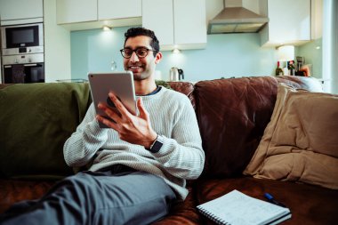 Evde çalışan, dijital tablette yazan, dinlenme odasındaki rahat koltukta oturan mutlu melez iş adamı.