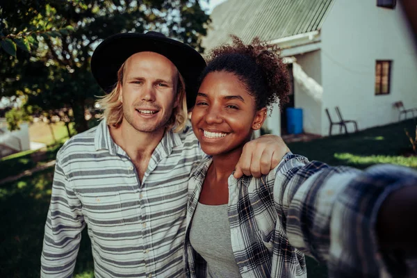 Gemengd ras paar glimlachen het nemen van selfie met cellulaire apparaat staan in de voorkant van witte boerderij — Stockfoto