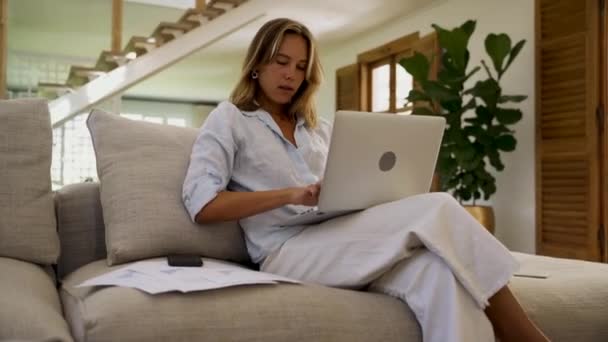 Blanke vrouw werken vanuit huis skimmen door papierwerk tijdens het typen op laptop — Stockvideo