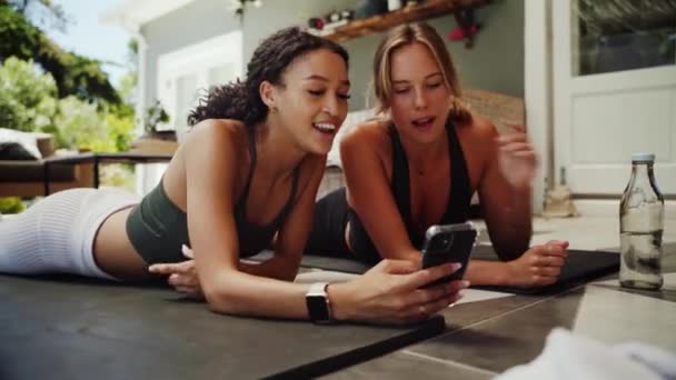 Amigos de carreras mixtas tumbados en la alfombra del gimnasio desplazándose en el dispositivo celular encontrar un buen entrenamiento para hacer en casa — Vídeo de stock