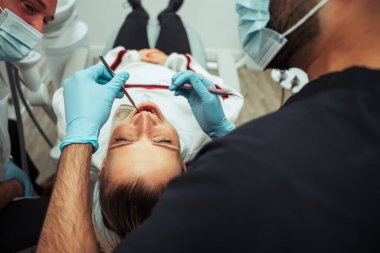 Beyaz kadın müşteri doktor yatağında yatarken erkek hemşire diş aletleriyle ameliyat ediyor.