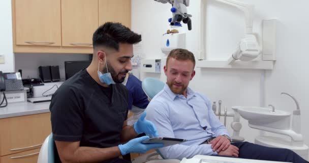Muž běloch sedí v křesle zubaře, zatímco ošetřovatel vysvětluje použití digitálního tabletu — Stock video