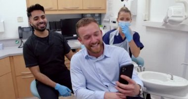 Beyaz erkek hasta, erkek ve kadın doktorlarla selfie çekiyor. Yeni temiz dişleri var.