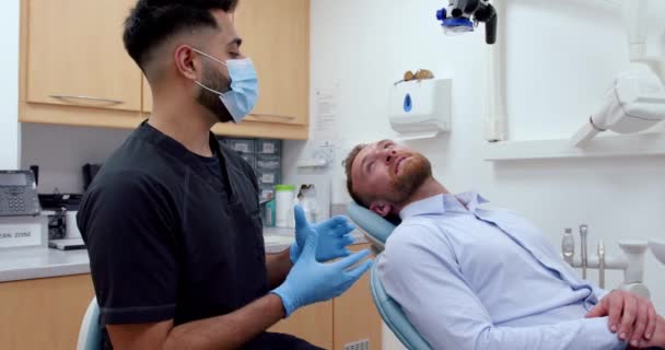 Male nurse wearing surgical mask chatting to male patient sitting in doctors office — Stockvideo