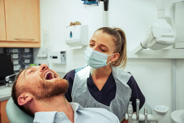 Καυκάσιος άνδρας ασθενής κάθεται στην καρέκλα ενώ έχουν τα δόντια καθαρίζονται από θηλυκό στοματικό υγιεινολόγο — Φωτογραφία Αρχείου