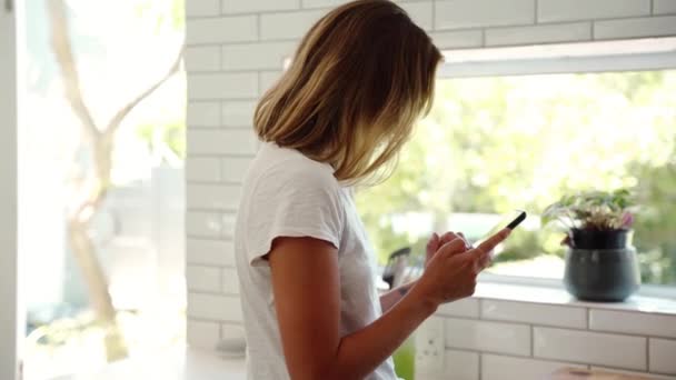 Femme caucasienne travaillant à partir de la maison tapant sur l'appareil cellulaire debout par la fenêtre dans la cuisine — Video