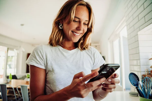 Studentessa caucasica che lavora da casa messaggiando sul dispositivo cellulare sorridente in piedi in cucina — Foto Stock