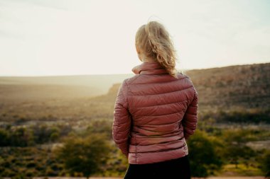 Dağın tepesinde yürüyüş yapan beyaz kadın elleri cebinde güneşin doğuşunu seyrediyor. 