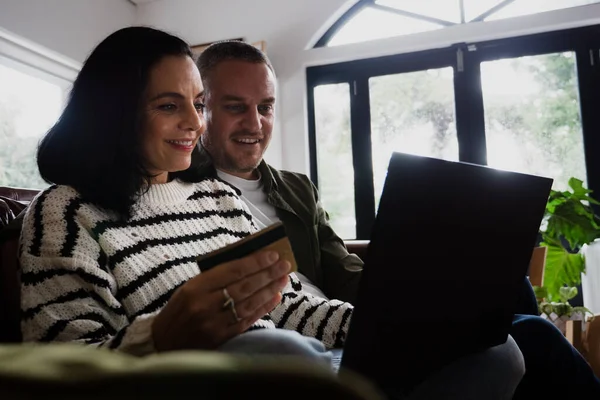 Boldog pár, hogy fizetési kártya gépelés a laptopon, miközben ül a kényelmes kanapén a nappaliban. — Stock Fotó