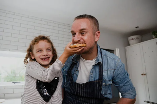 Filha rindo enquanto alimentava o pai sanduíche caseiro em pé na cozinha. — Fotografia de Stock