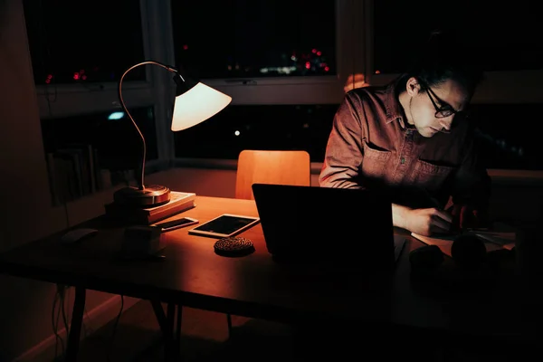 Caucasian business man working late at night organising week plans sitting writing in notepad and laptop