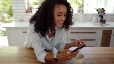 Karışık ırk kadınlarıyla afro saçlı kadınlar mutfakta oturmuş, gülümserken hücresel cihazlarla yazı yazıyorlar. 