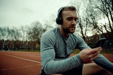 Kafkas erkek sporcu izleme sahasında oturuyor kulaklık takıyor ve elinde cep telefonu var. 