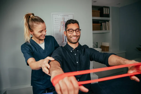 Blanke vrouwelijke fysiotherapeut assisteren mannelijke patiënt houden weerstand band — Stockfoto