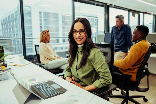 İş arkadaşları toplantı yaparken Hintli iş kadını ofiste dizüstü bilgisayarının arkasında oturmuş gülümsüyor.