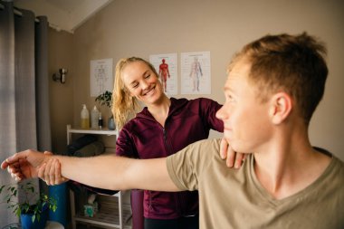 Kadın fizyoterapist erkek hastanın kolunu gererken gülümsüyor ve pilates stüdyosunda oturuyor.