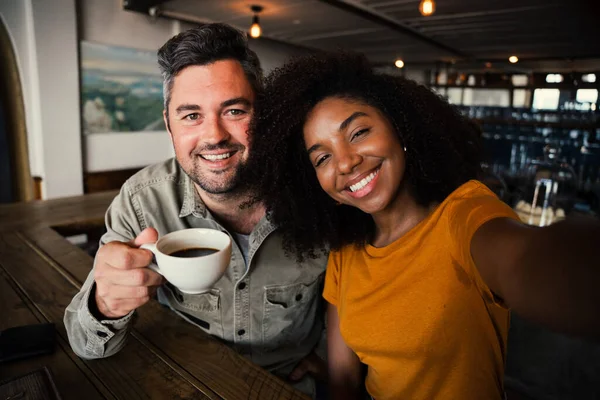 Pareja de raza mixta sonriendo maravillosamente para selfie tomada en el teléfono inteligente mientras bebe café caliente en la cafetería de moda — Foto de Stock