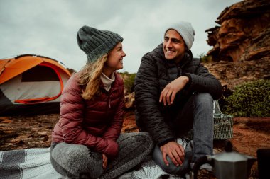 Dağda kamp yaparken sohbet eden beyaz tenli romantik çift gülümsüyor.