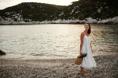 Beyaz kadın sahilde sandaletle yürüyor.