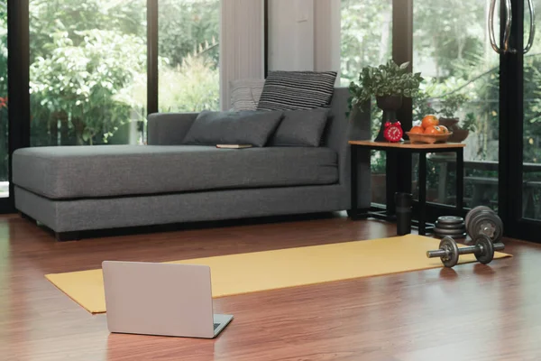 Laptop, Yellow yoga mat and dumbbell on the floor in modern living room for exercise at home.