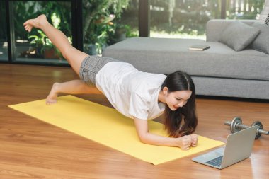 Günlük kıyafet giymiş Asyalı bir kadın evdeki bilgisayar laptopunda online antrenman dersleri izleyerek egzersiz öğreniyor..