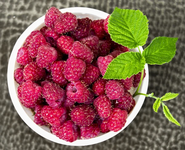 På bordet det är en liten vit tallrik fylld med färska saftiga bär mogna röda hallon och dekorerad med ett grönt blad av hallon bush — 图库照片