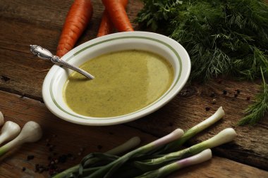 Bir seramik kase, havuç, soğan, sarımsak ve ahşap masa üzerinde otlar içinde yeşil bezelye çorbası