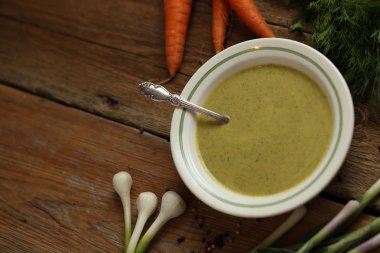 Bir seramik kase, havuç, soğan, sarımsak ve ahşap masa üzerinde otlar içinde yeşil bezelye çorbası