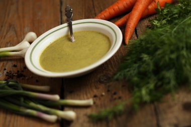 Bir seramik kase, havuç, soğan, sarımsak ve ahşap masa üzerinde otlar içinde yeşil bezelye çorbası