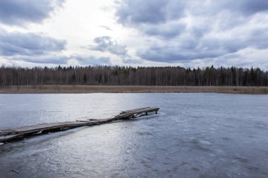 Uzun ahşap iskeleli donmuş bir göl manzarası. Göletin uzak tarafındaki bulutlu gökyüzü ve orman.