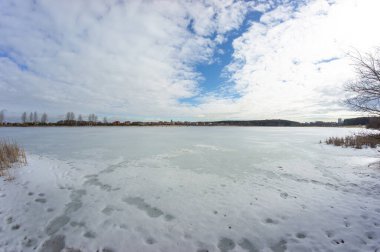 Baharın başında donmuş bir göl manzarası. Balıkçılar gölün ortasındaki buzun üzerinde oturuyorlar. Uzak kıyıda bir köy görünüyor..