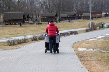 Çifte bebek arabası olan bir baba baharın başında soğuk ve ıssız bir şehir parkında gezinir. Parlak kırmızı ceketli bir adam..